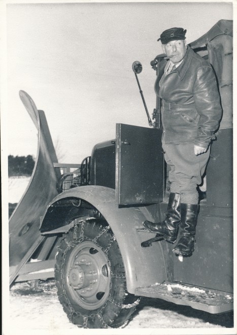 Standing on side of snow plough with cigar in mouth. Chains wrapped around tyres for extra grip. 1966. 